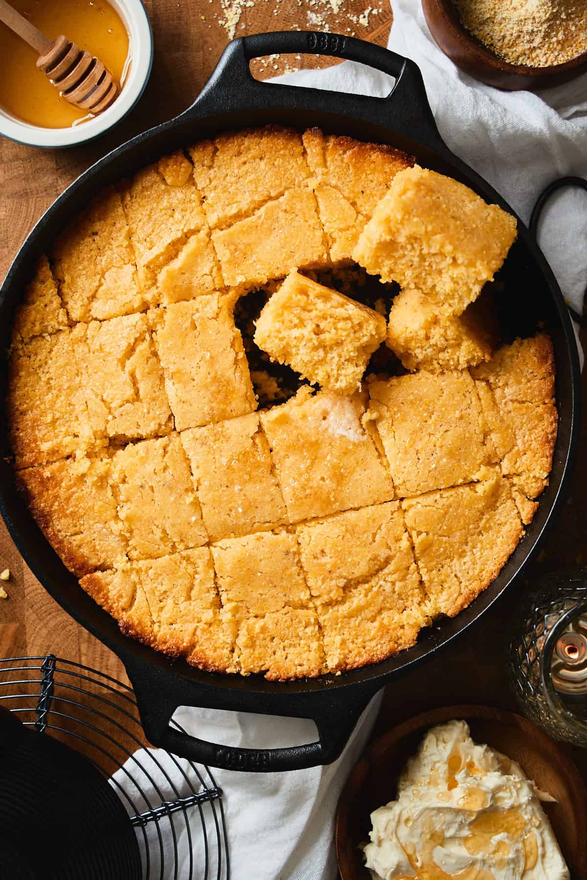 Squares of cornbread cut from a skillet, with some facing up toward the camera, surrounded by whipped honey butter, candles, ingredients, and a linen napkin.