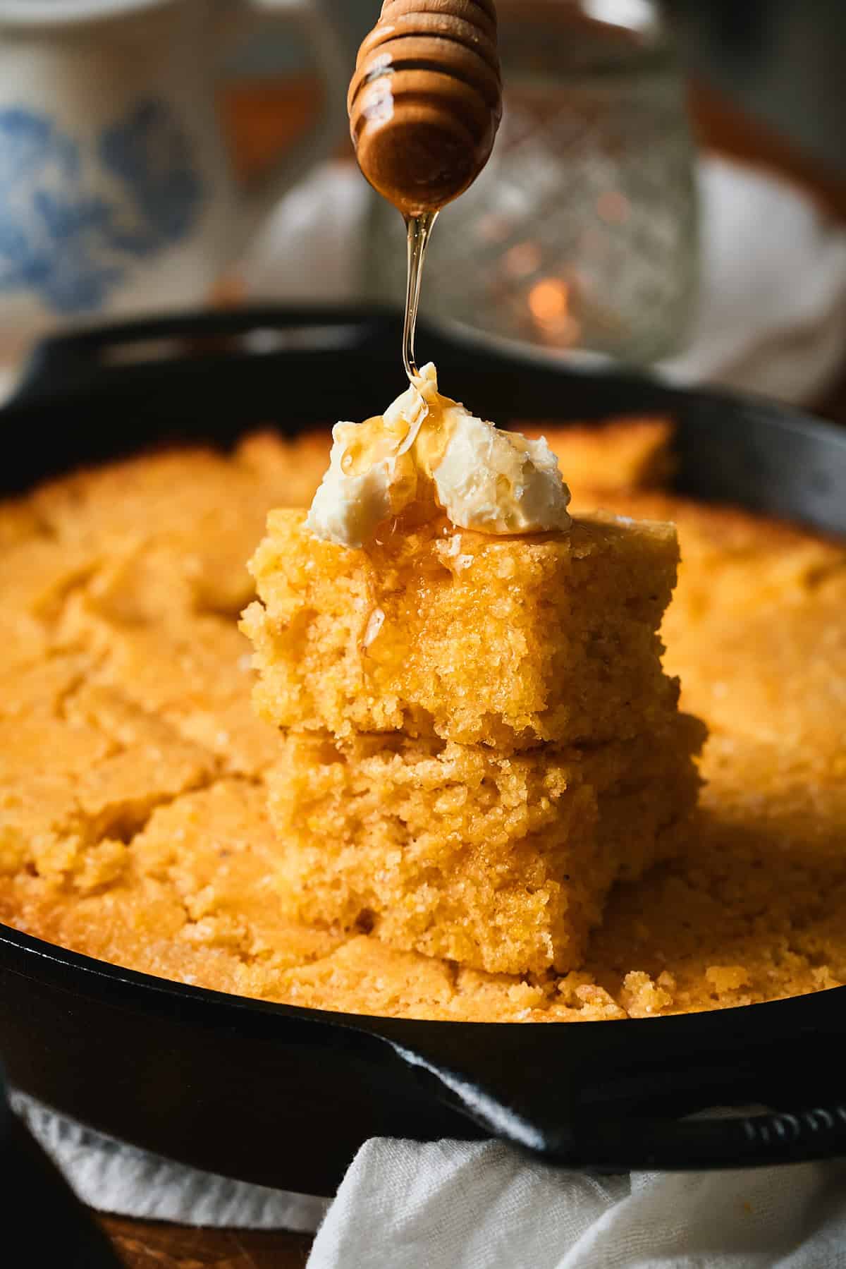Photo of two pieces of cornbread stacked in cast iron skillet and topped with whipped butter while being drizzled with honey.