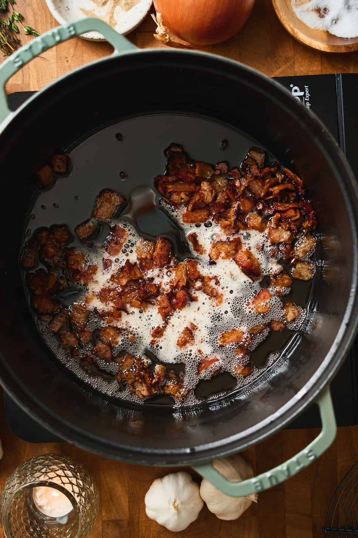 Crispy bacon pieces in rendered fat in a green Staub Dutch oven.