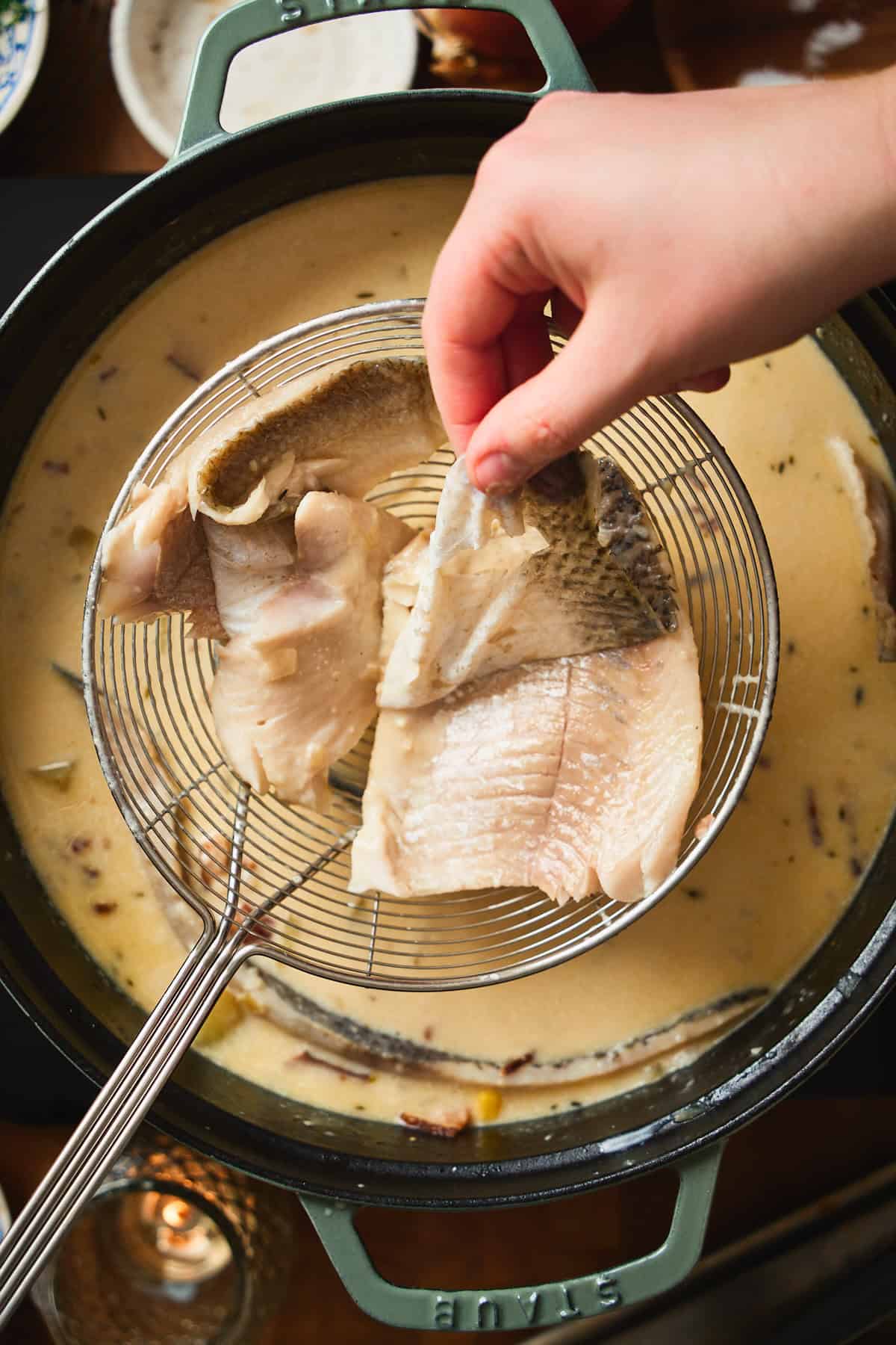 Hand peeling skin off a poached whitefish fillet resting on a spider strainer.