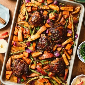 Overhead square shot of a sheet pan of glazed chicken thighs with roasted apple wedges, sweet potatoes, green beans, and red onion, garnished with fresh herbs.