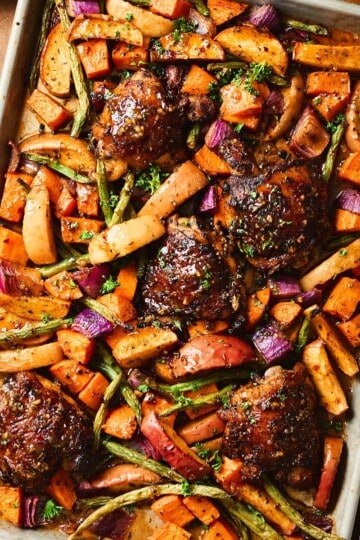 Overhead square shot of a sheet pan of glazed chicken thighs with roasted apple wedges, sweet potatoes, green beans, and red onion, garnished with fresh herbs.