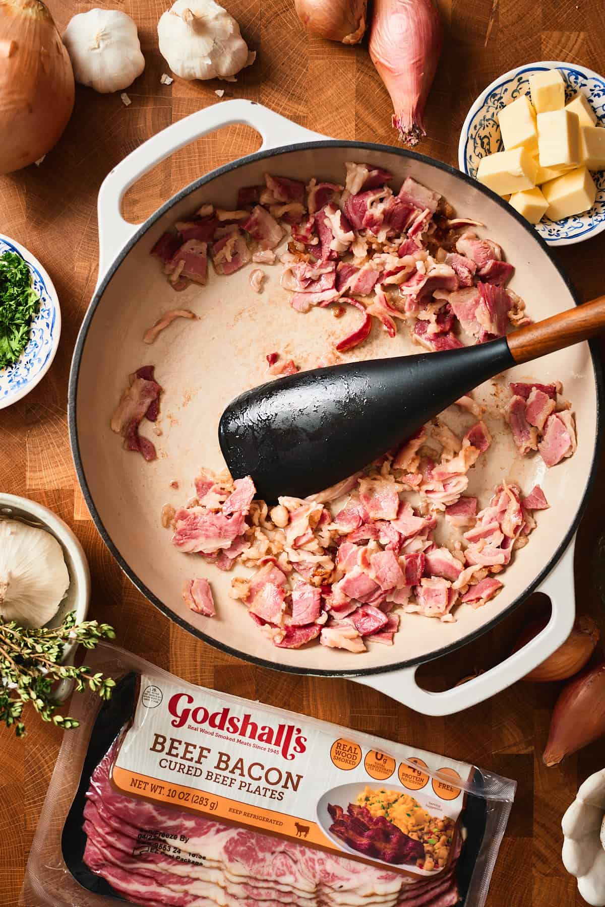 Raw bacon pieces added to a white skillet surrounded by fresh ingredients.