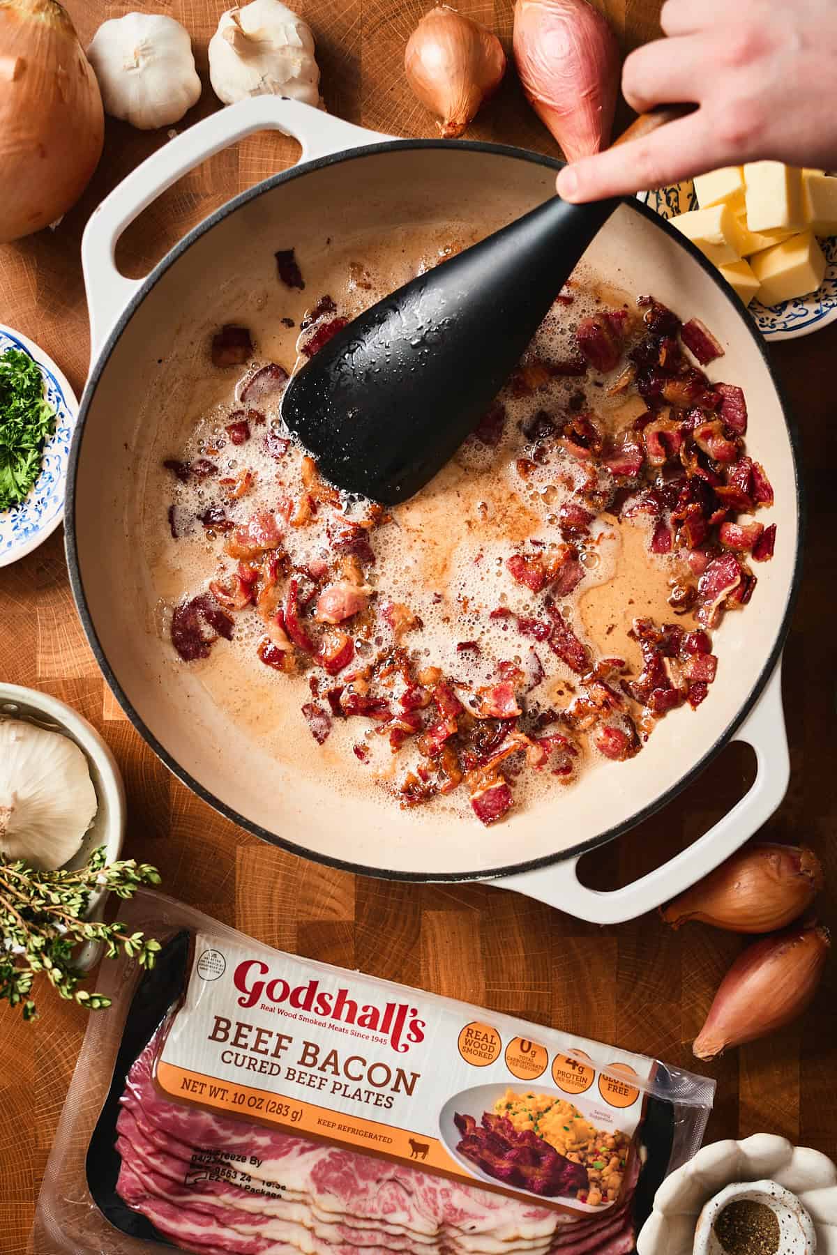 Cooked Godshall's beef bacon pieces in a white skillet surrounded by fresh ingredients,