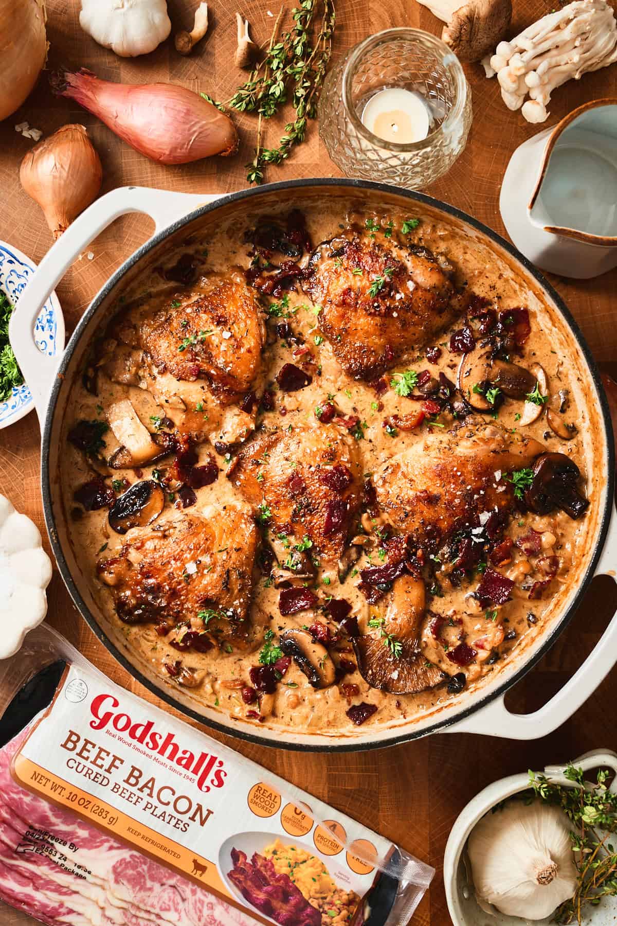 Creamy mushroom chicken thighs in a white skillet with Godshall's beef bacon package, overhead view.