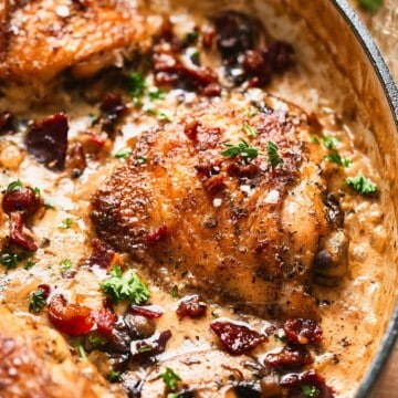 Close-up of a creamy mushroom chicken thigh with crispy beef bacon and fresh herbs in a rich cream sauce in a skillet.