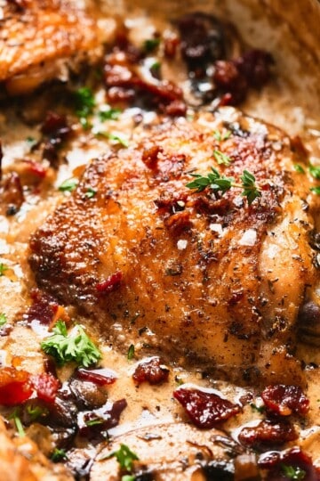 Close-up of a creamy mushroom chicken thigh with crispy beef bacon and fresh herbs in a rich cream sauce in a skillet.
