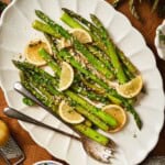 Overhead close-up of lemon garlic asparagus on a white scalloped platter with lemon slices, parsley, and serving utensils.