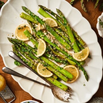 Overhead close-up of lemon garlic asparagus on a white scalloped platter with lemon slices, parsley, and serving utensils.