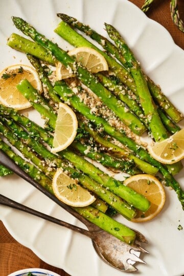 Overhead close-up of lemon garlic asparagus on a white scalloped platter with lemon slices, parsley, and serving utensils.