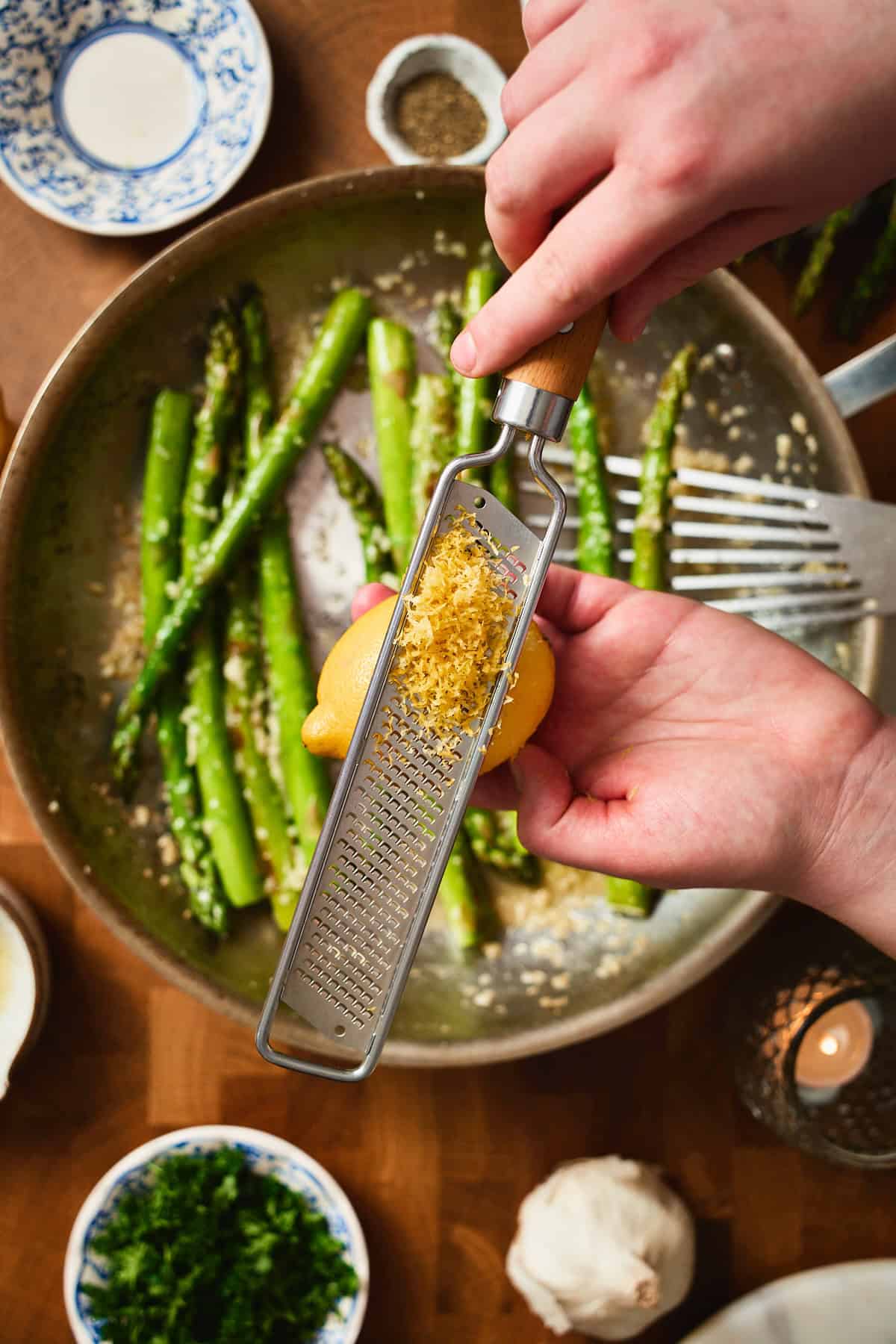 Hands zesting a lemon over pan fried asparagus in a skillet.