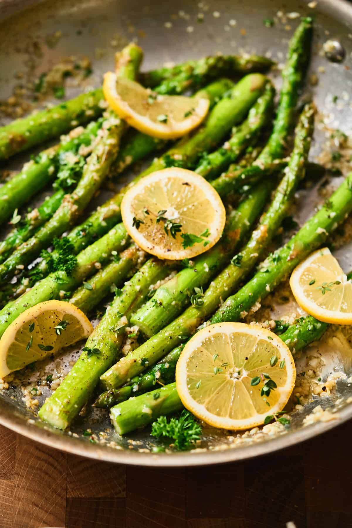 Hero Close-up of pan fried asparagus with lemon garlic butter in a skillet, garnished with lemon slices, parsley, and thyme.