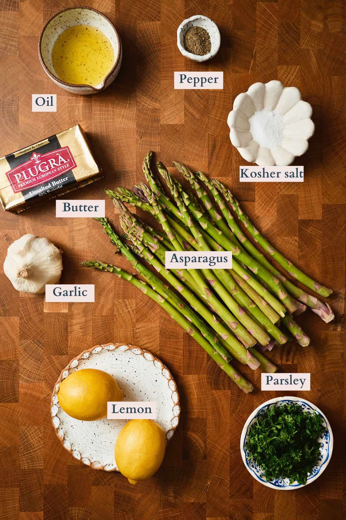 Labeled ingredients for pan fried asparagus with lemon garlic butter on a wooden board, including asparagus, butter, garlic, lemon, oil, parsley, salt, and pepper.