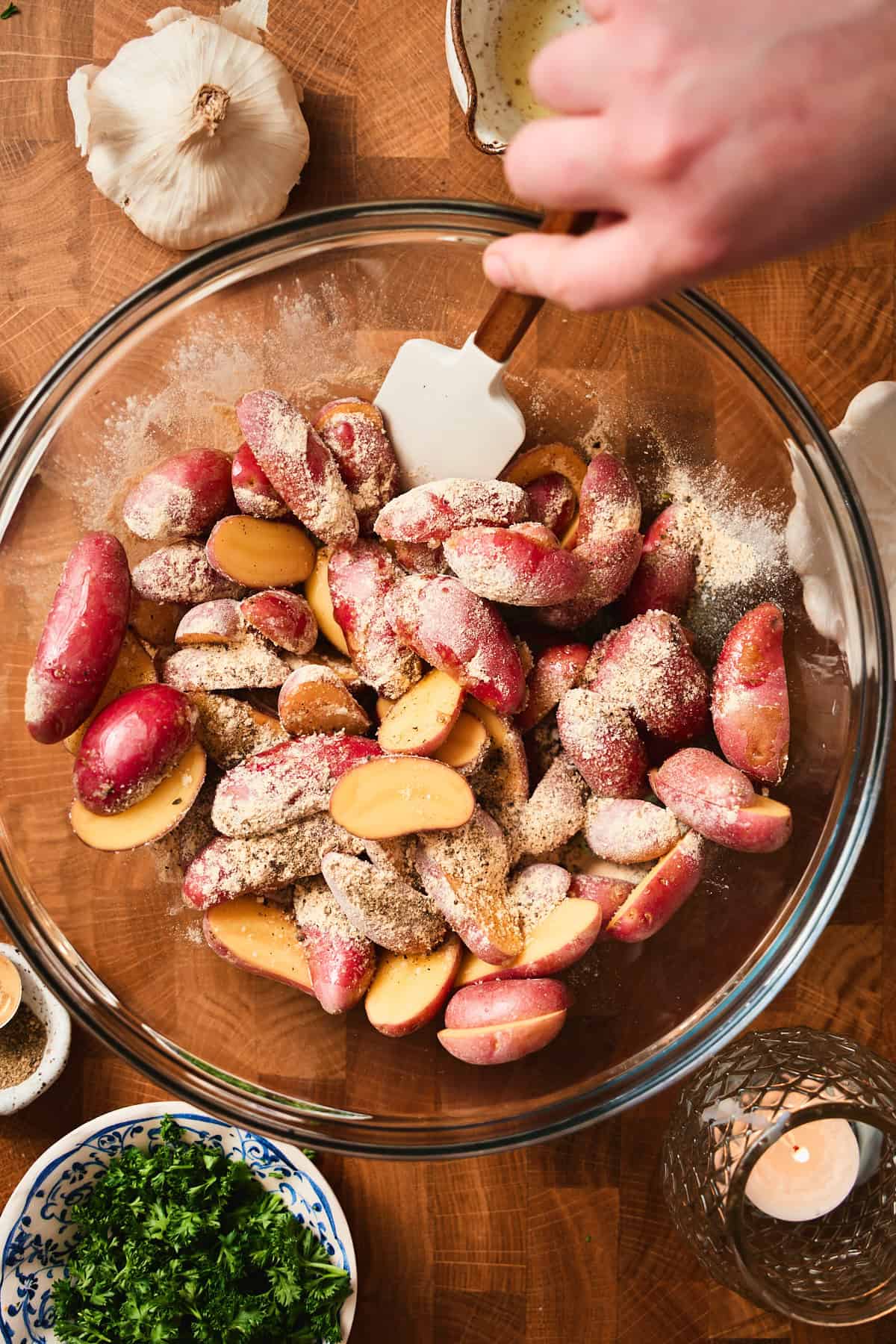 Seasoned potatoes in a glass bowl with a hand using a rubber spatula to toss seasoning on the potatoes.