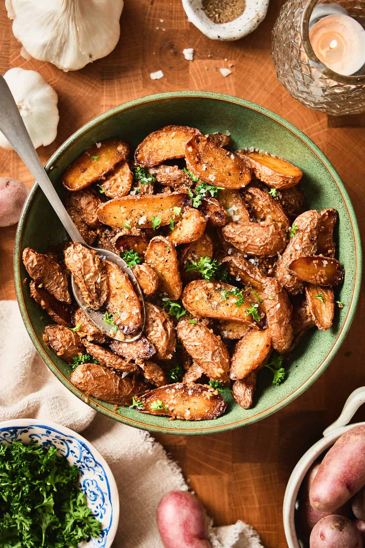 Overhead bowl of garlic butter roasted baby potatoes with parsley, flaky salt, garlic, and raw potatoes on a wooden surface.