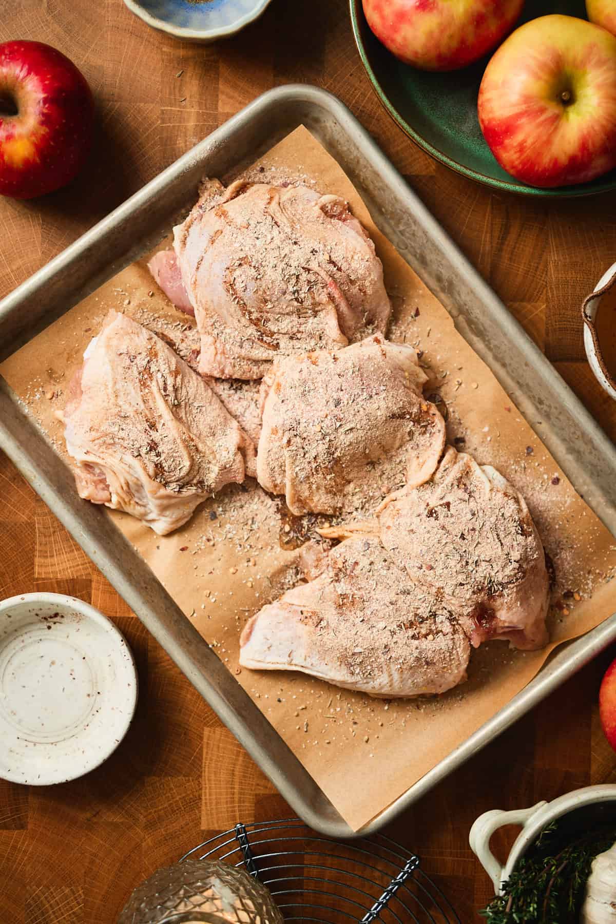 Raw bone-in chicken thighs coated in a dry spice rub on a parchment-lined sheet pan, surrounded by fresh apples.