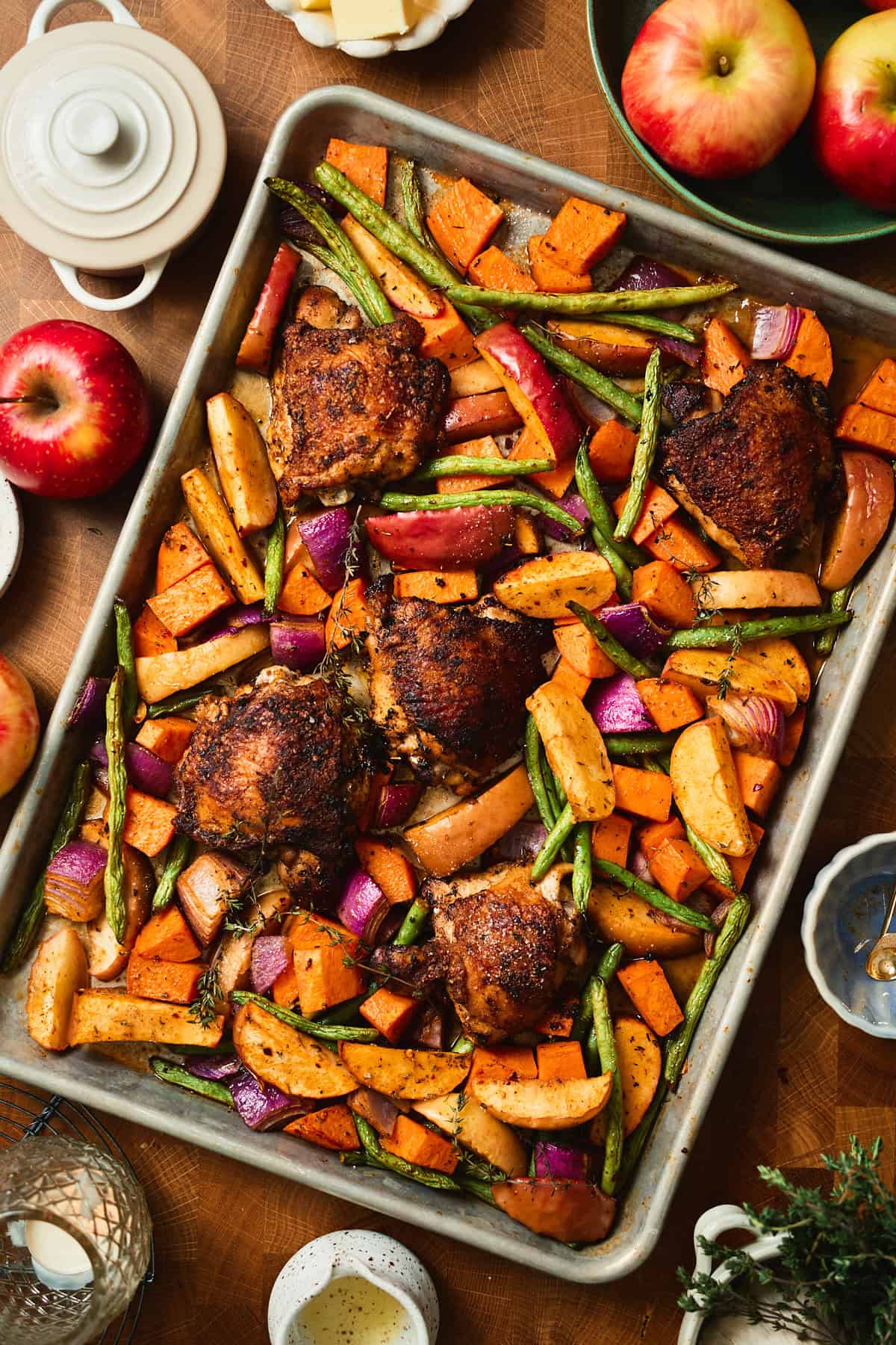 Sheet pan of seasoned chicken thighs nestled among apple wedges, sweet potatoes, green beans, and red onion before glazing.