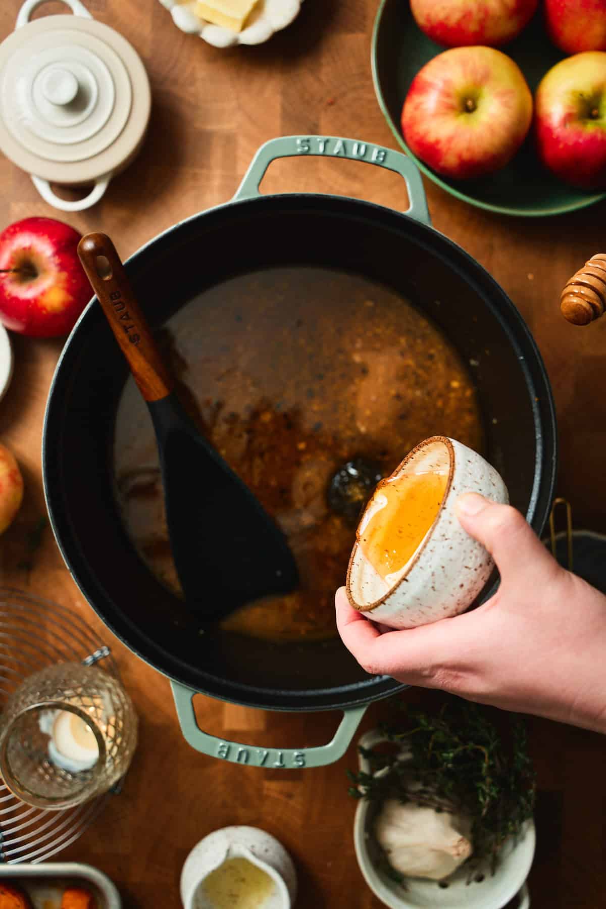 Hand pouring honey into a Staub Dutch oven with reduced apple cider glaze.