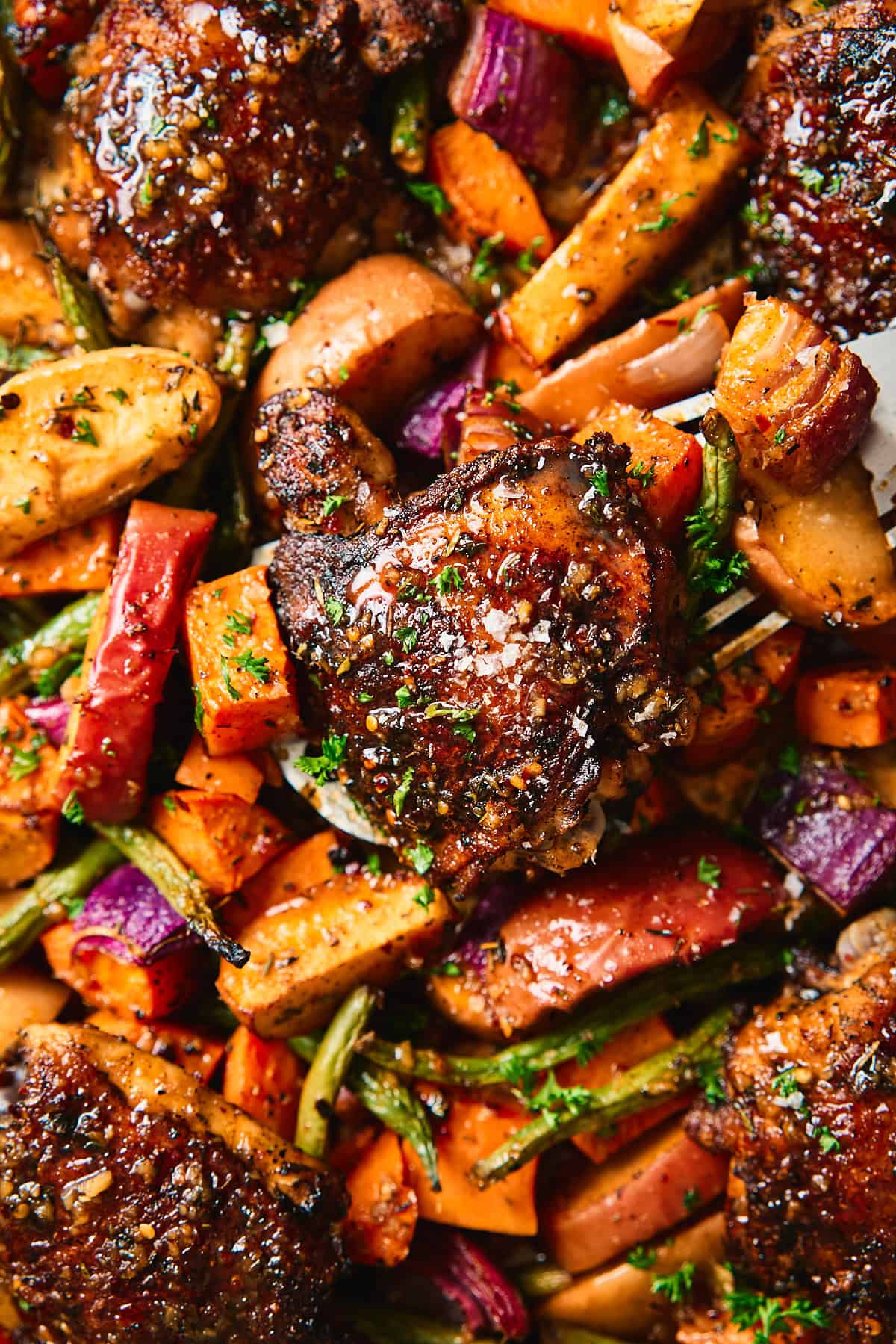 Close-up of a glazed chicken thigh being lifted from a sheet pan surrounded by caramelized sweet potatoes, apples, red onion, and green beans.