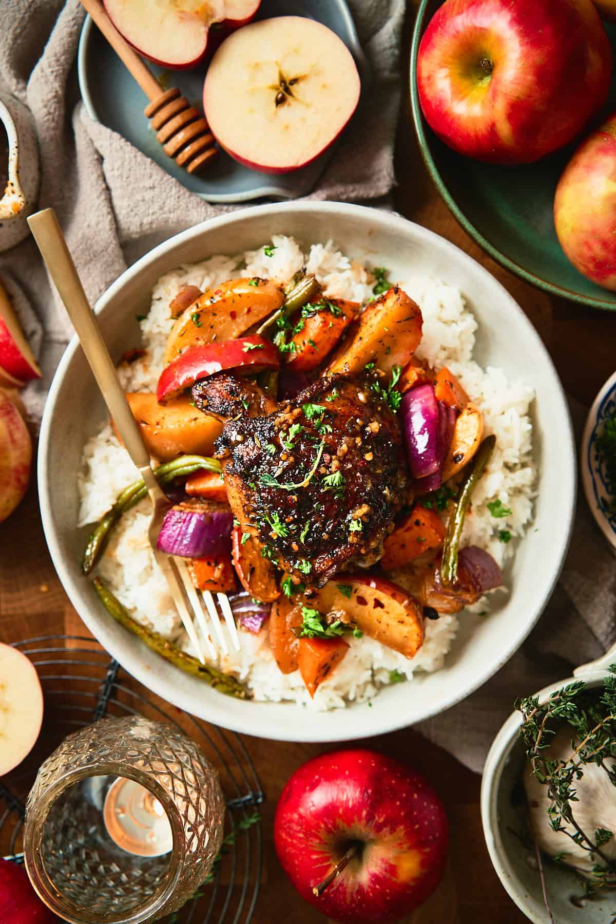 Overhead bowl of glazed chicken thigh with roasted apples, sweet potatoes, red onion, and green beans over white rice, garnished with fresh herbs.