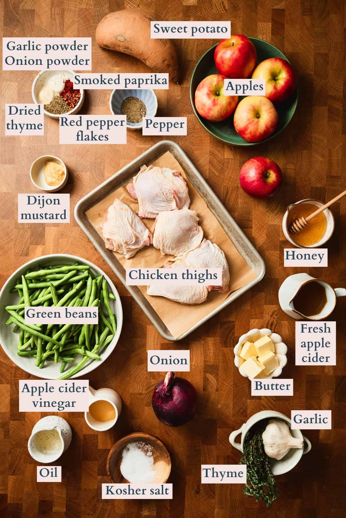 Labeled ingredients for sheet pan chicken and apples laid out on a wooden surface, including chicken thighs, apples, sweet potato, green beans, butter, honey, apple cider, and spices.