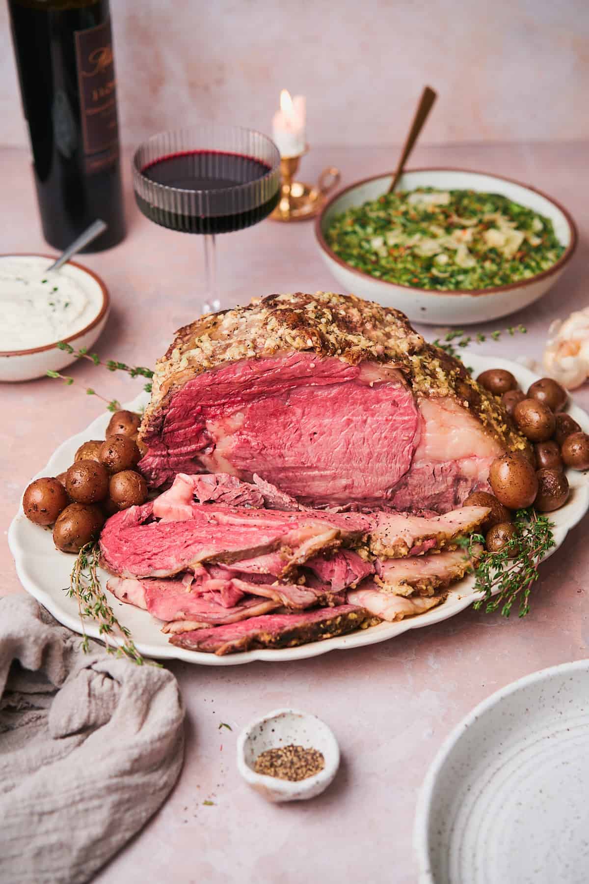 Sliced slow cooker prime rib roast on a white scalloped platter with roasted potatoes, fresh thyme, and a glass of red wine.