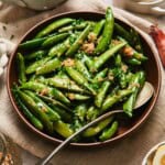Close-up of buttery sautéed sugar snap peas with shallots and parsley in a brown bowl.