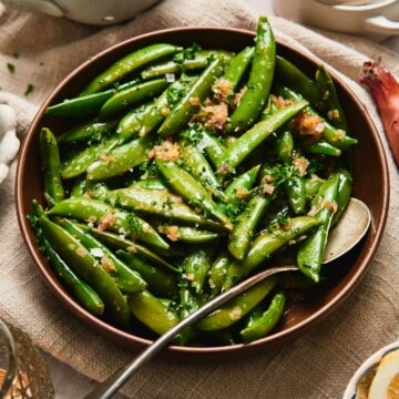 Close-up of buttery sautéed sugar snap peas with shallots and parsley in a brown bowl.