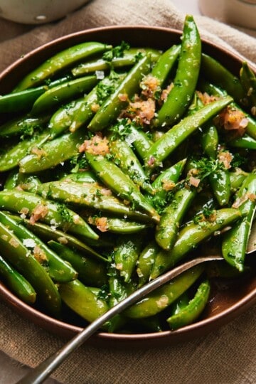 Close-up of buttery sautéed sugar snap peas with shallots and parsley in a brown bowl.