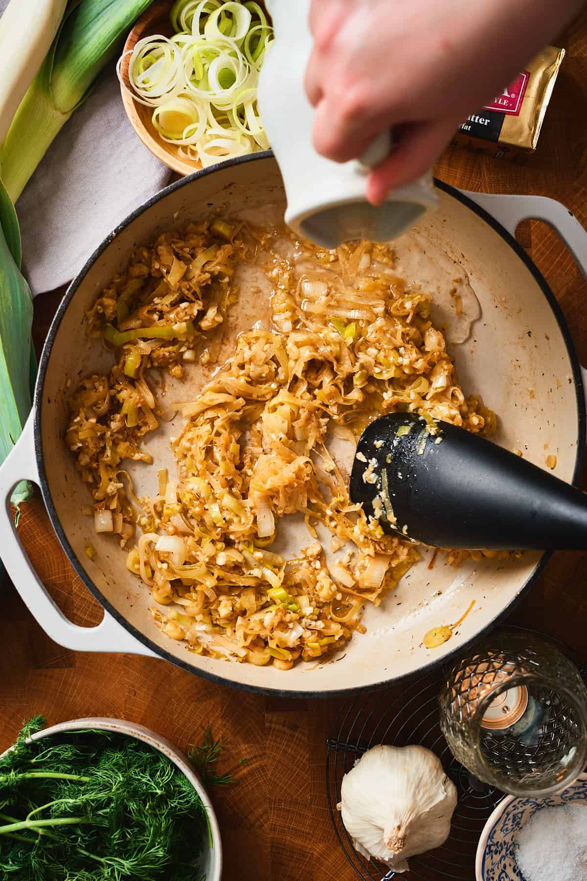 Hand pouring white wine into a pan of caramelized leeks and garlic.