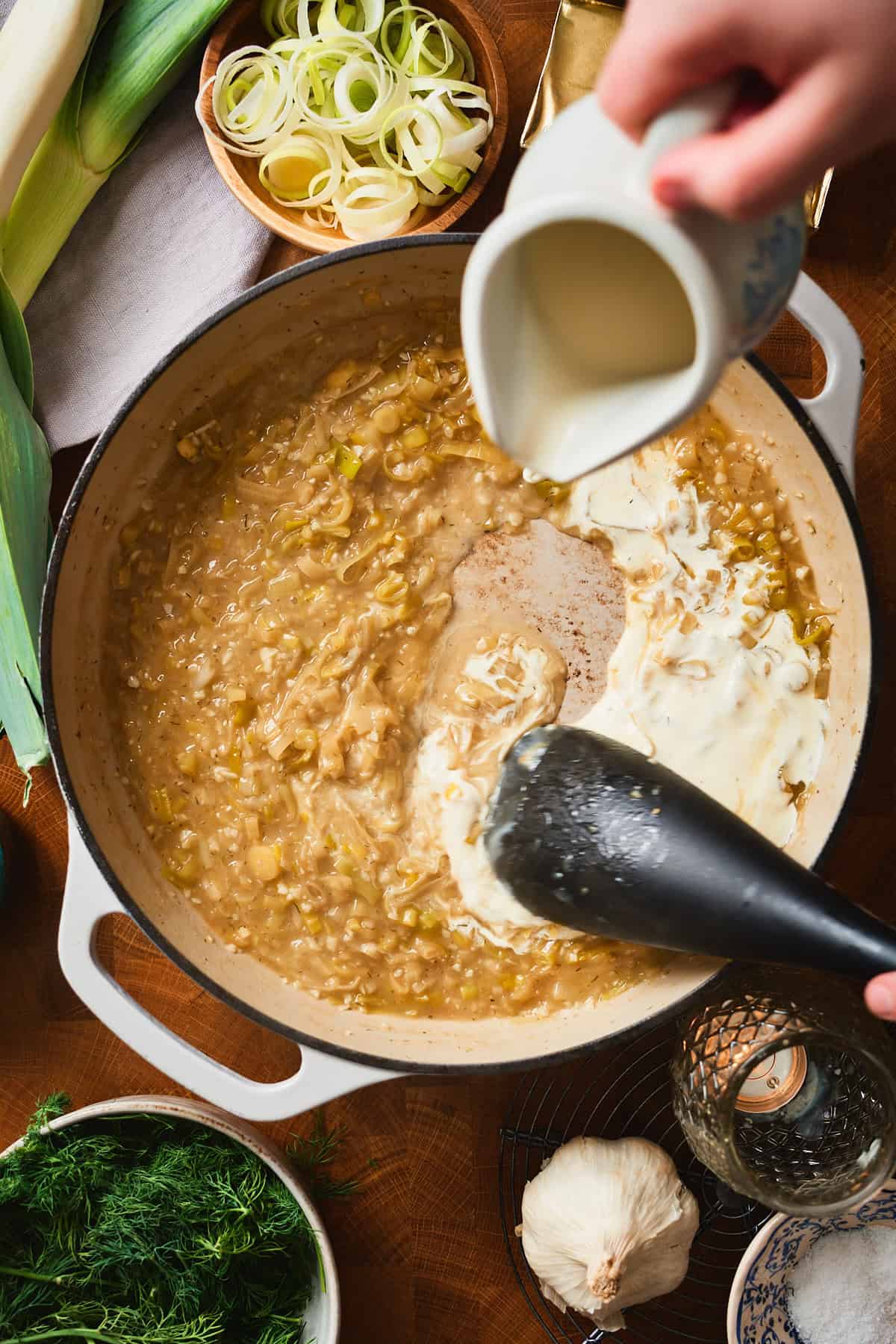 Hand pouring heavy cream into a simmering leek sauce.