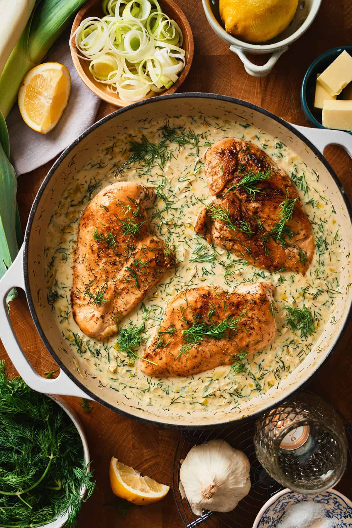 Overhead view of creamy leek chicken with dill sauce in a white pan surrounded by fresh leeks, dill, lemon, and garlic.