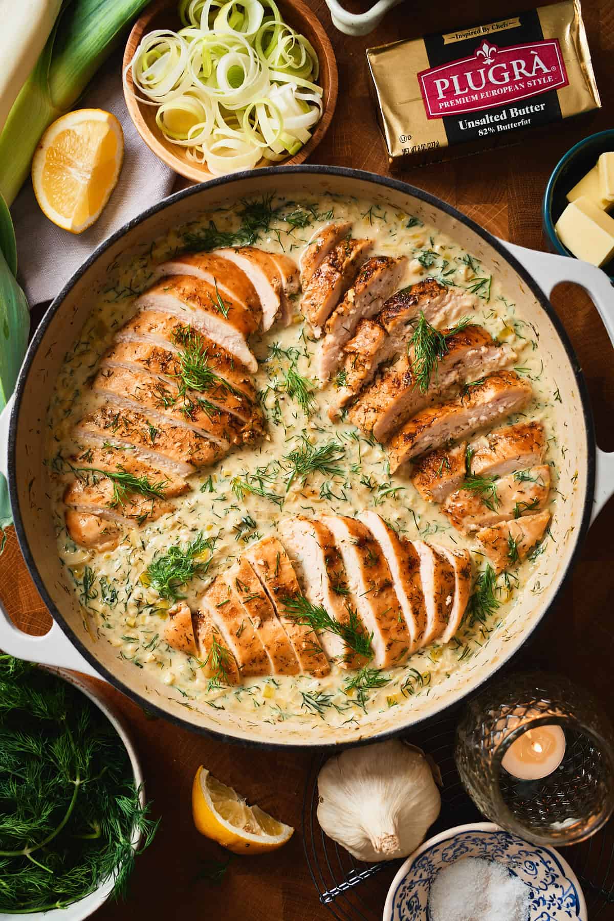 Overhead shot of sliced chicken breasts in creamy leek dill sauce in a skillet, with a Plugrà butter package, lemons, leeks, and garlic nearby.