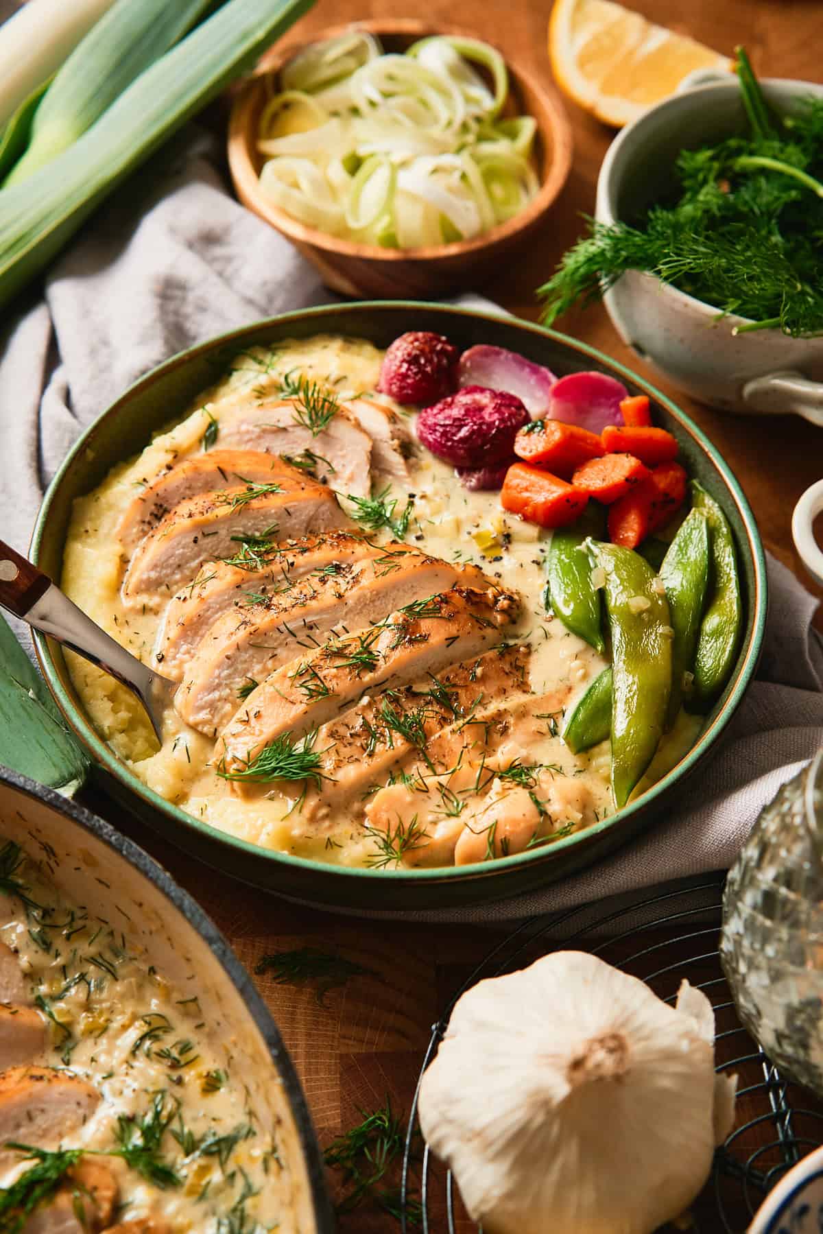 Close-up of sliced creamy leek chicken in lemon dill cream sauce over mashed potatoes with snap peas and carrots in a green bowl.