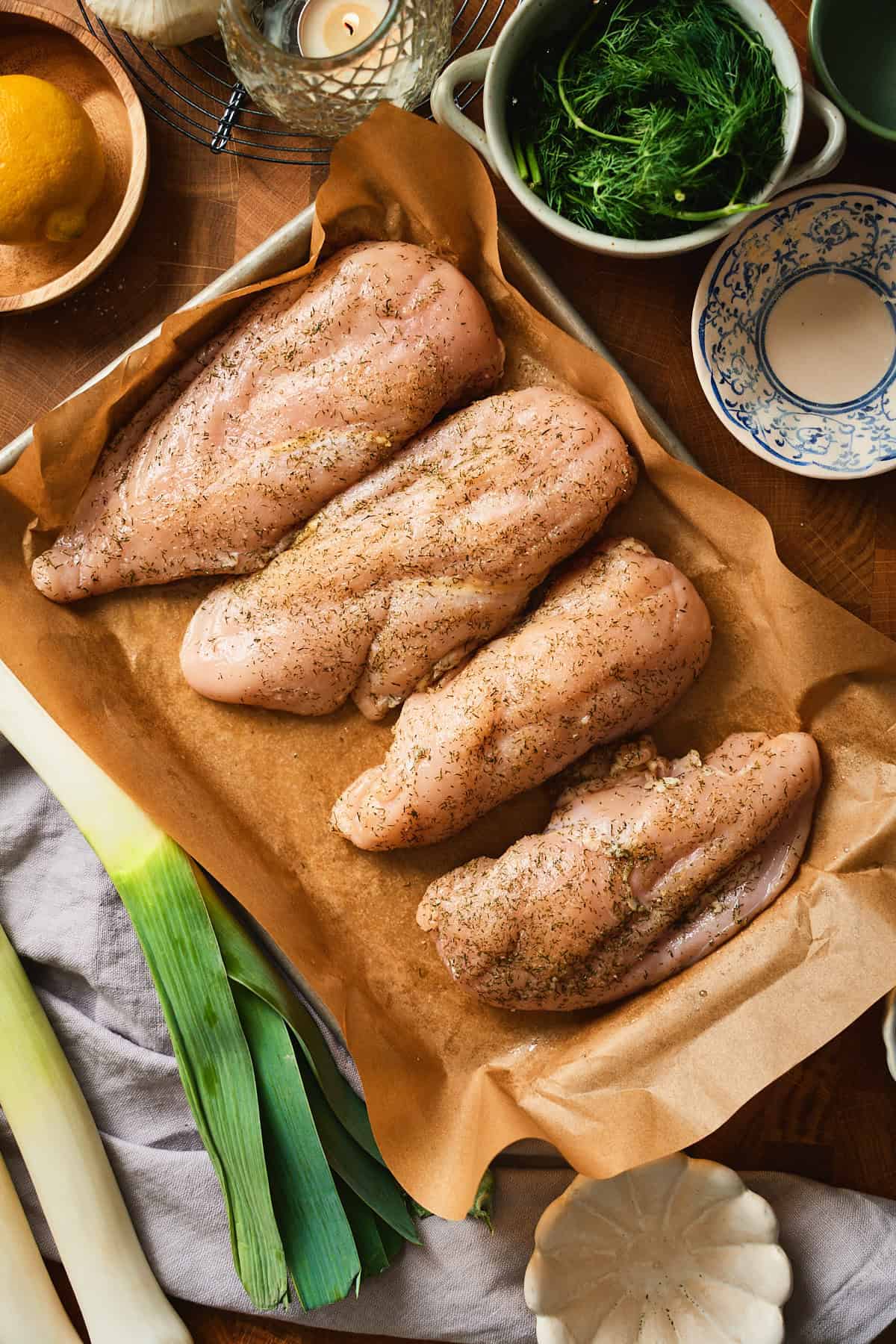Four seasoned raw chicken breasts on parchment paper surrounded by fresh leeks, dill, and lemon.
