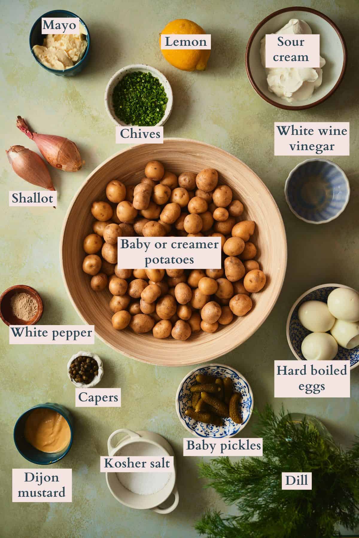 Labeled overhead flat lay of ingredients for Swedish dill potato salad - baby creamer potatoes, sour cream, mayo, Dijon mustard, white wine vinegar, lemon, shallot, baby pickles, capers, fresh dill, chives, hard-boiled eggs, white pepper, and kosher salt.