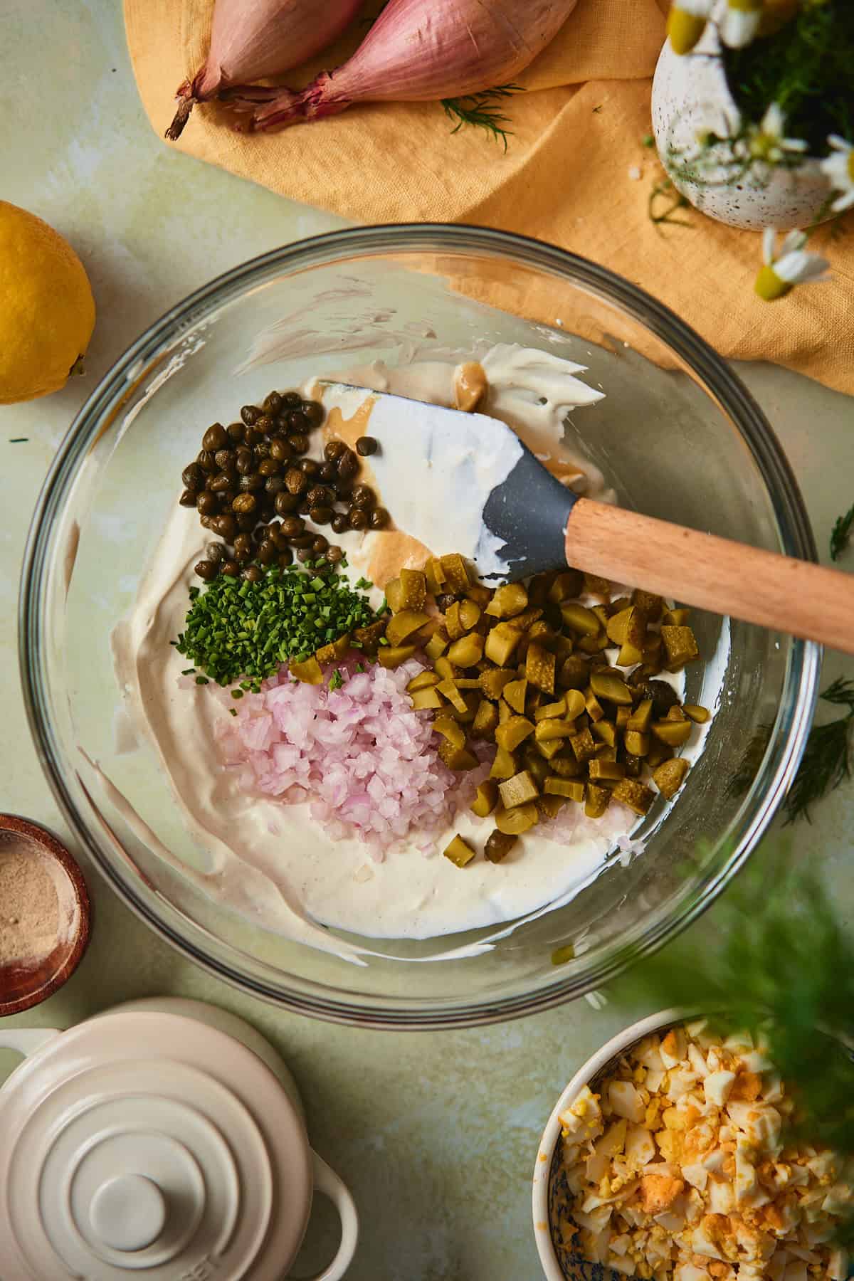 Chopped baby pickles, capers, minced shallot, and chives arranged on top of sour cream dressing in a glass bowl.