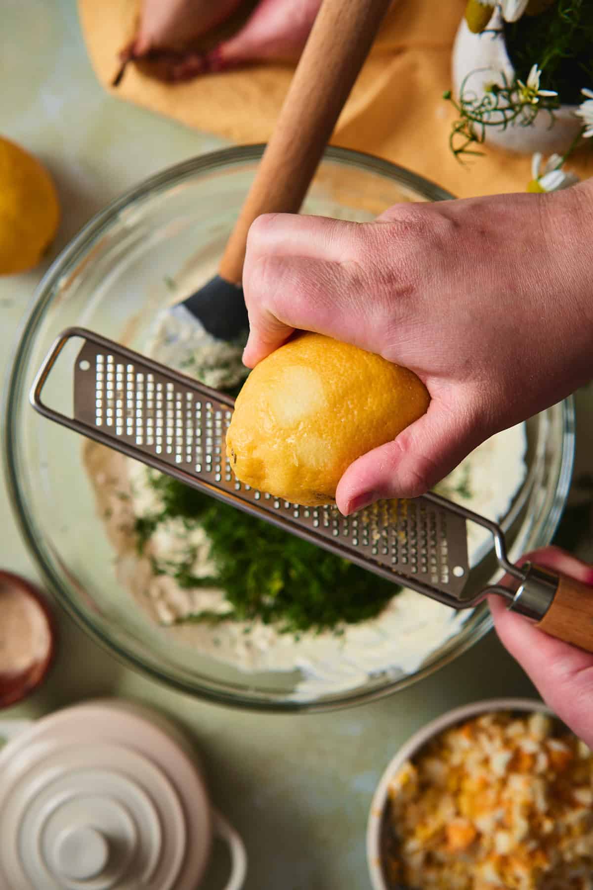 Hands zesting a fresh lemon over a bowl of dill potato salad dressing using a microplane.