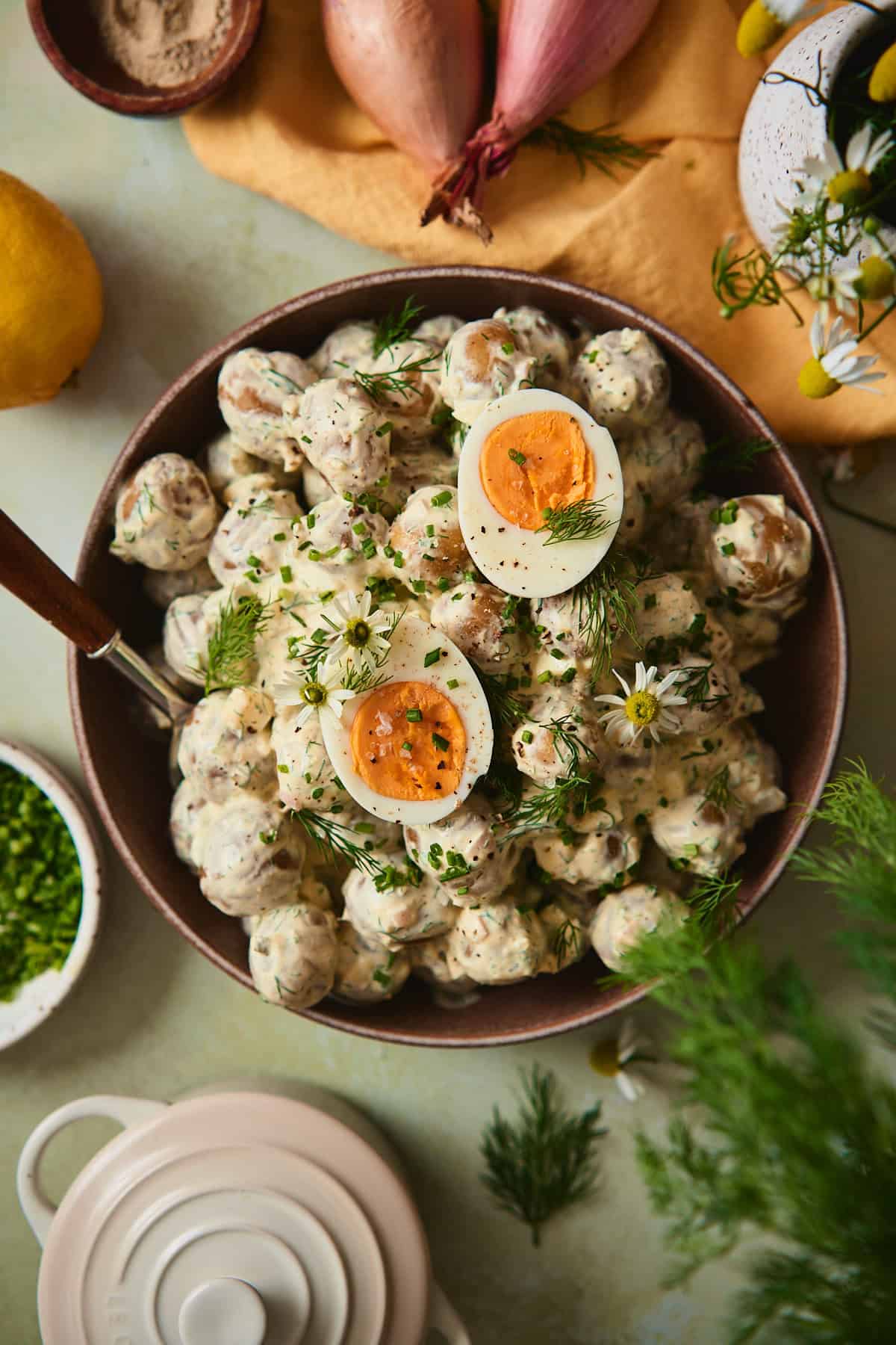 Overhead view of creamy Swedish dill potato salad in a brown ceramic bowl, topped with halved hard-boiled eggs, fresh dill, chives, and daisies on a mustard yellow linen.