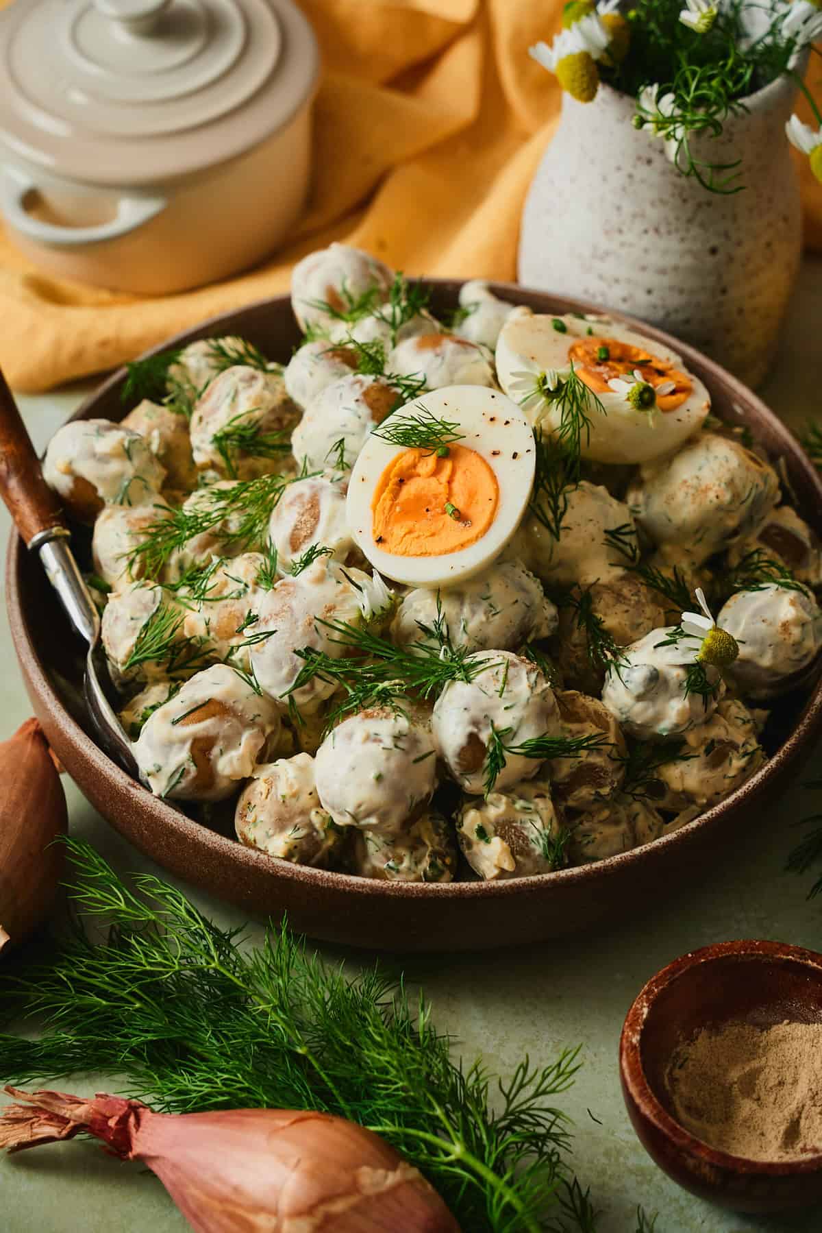 Side angle of creamy Swedish dill potato salad in a brown ceramic bowl with halved hard-boiled eggs, fresh dill, daisies, and a wooden serving spoon.