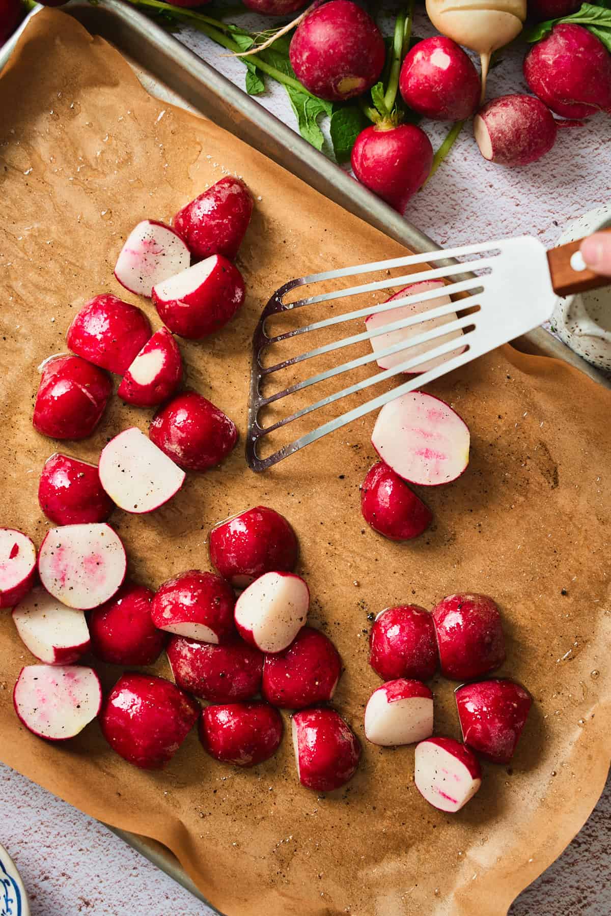 Halved raw radishes being arranged cut-side-down on a parchment-lined baking sheet with a spatula.