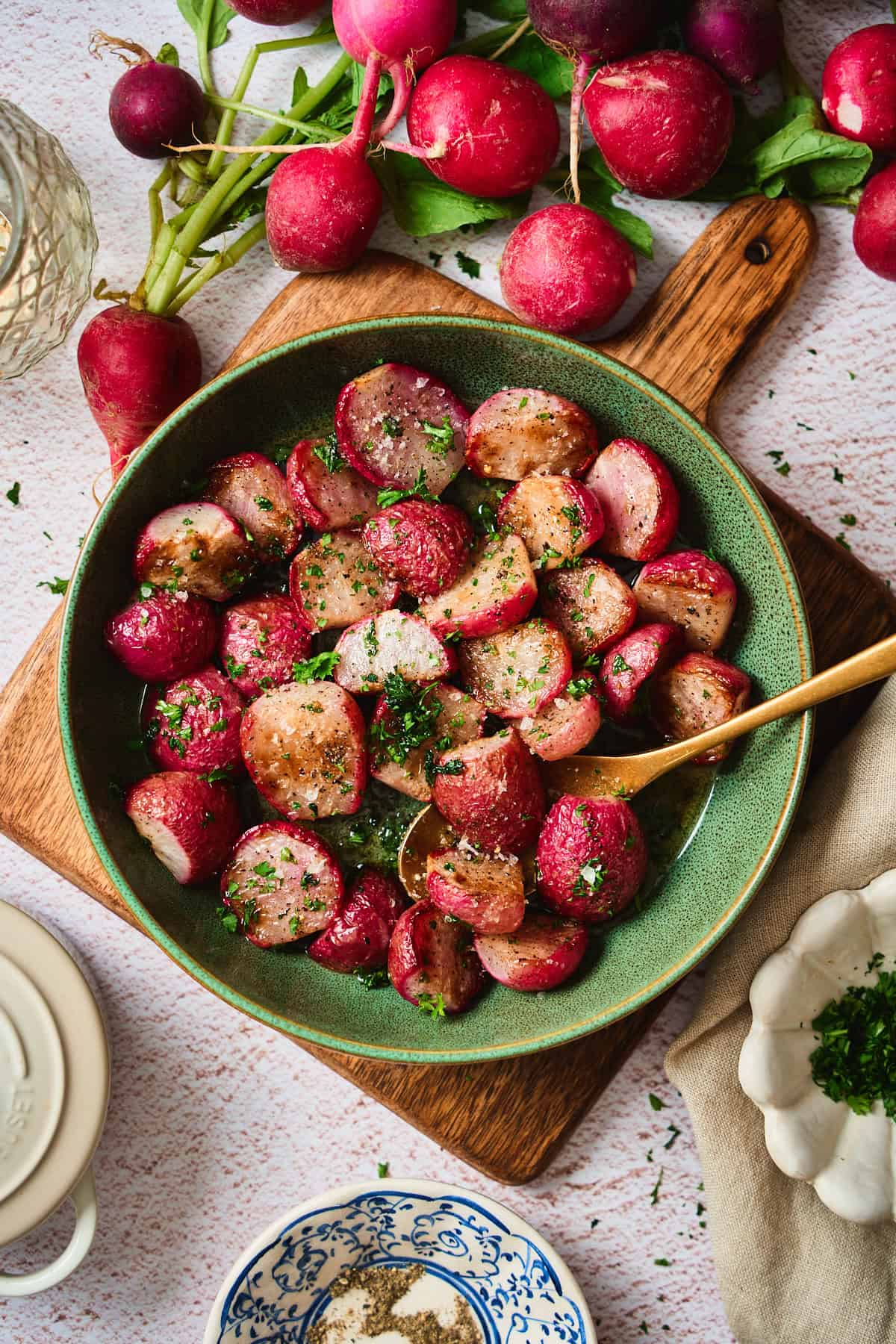 Roasted radishes with brown butter in a green bowl on a wood board, surrounded by whole radishes and a lit lantern.