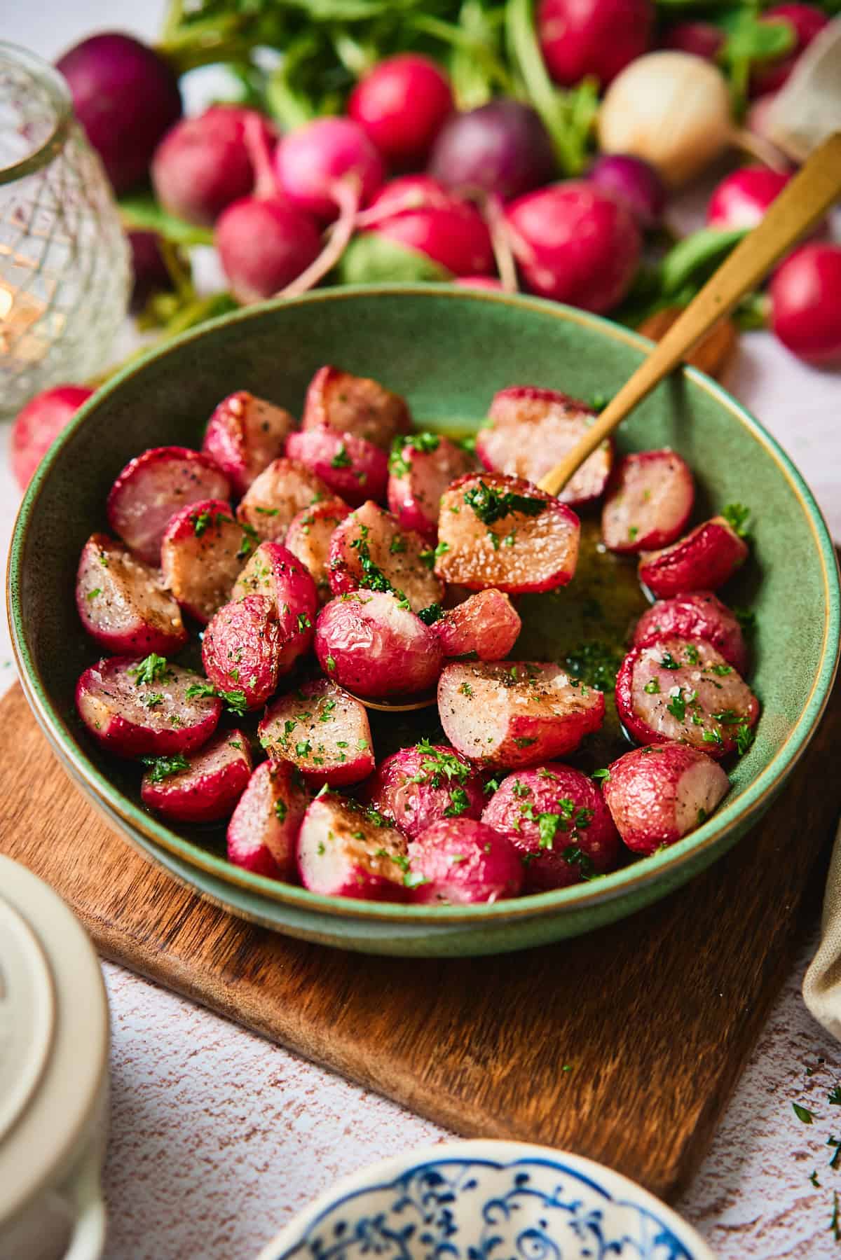 Roasted radishes with brown butter in a green bowl on a wood board, surrounded by whole radishes and a lit lantern.