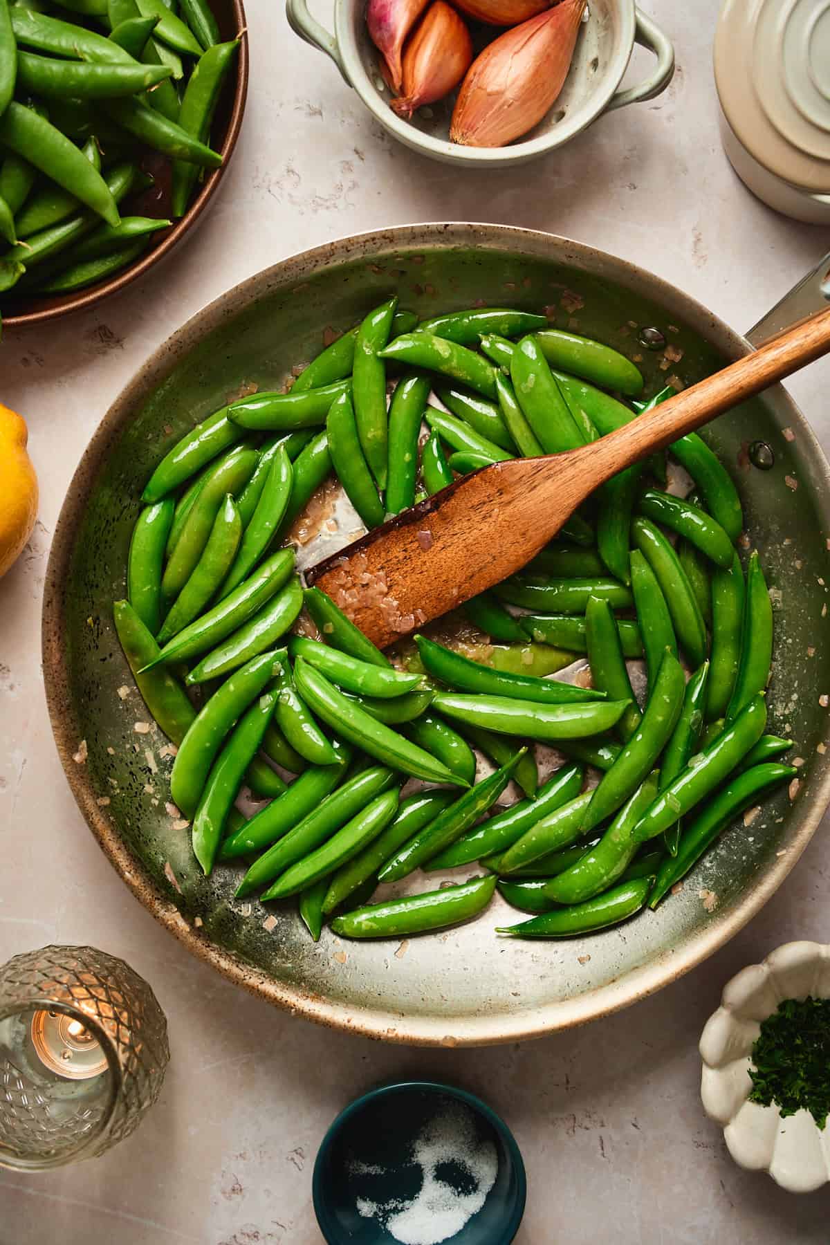 Bright green snap peas being tossed in a buttery skillet with a wooden spoon.
