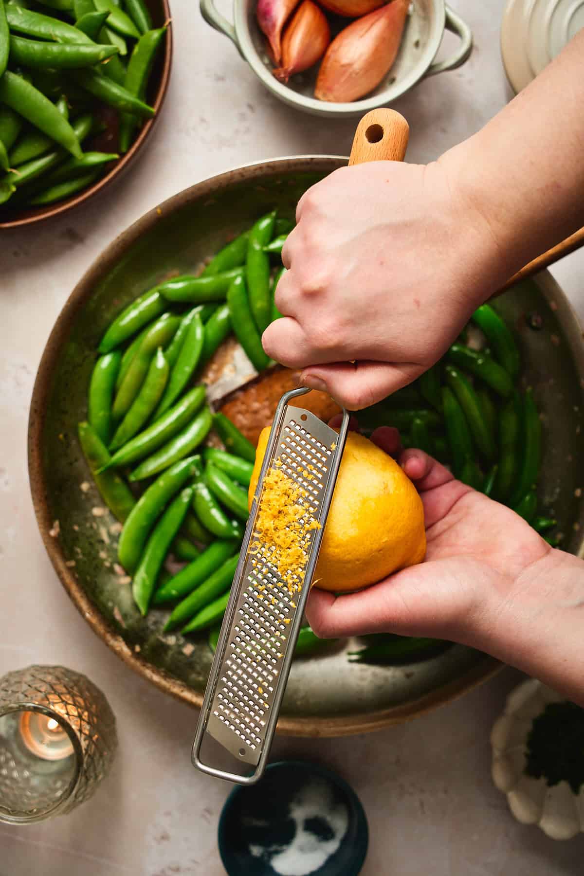 Hands zesting a lemon directly over a skillet of sautéed snap peas.