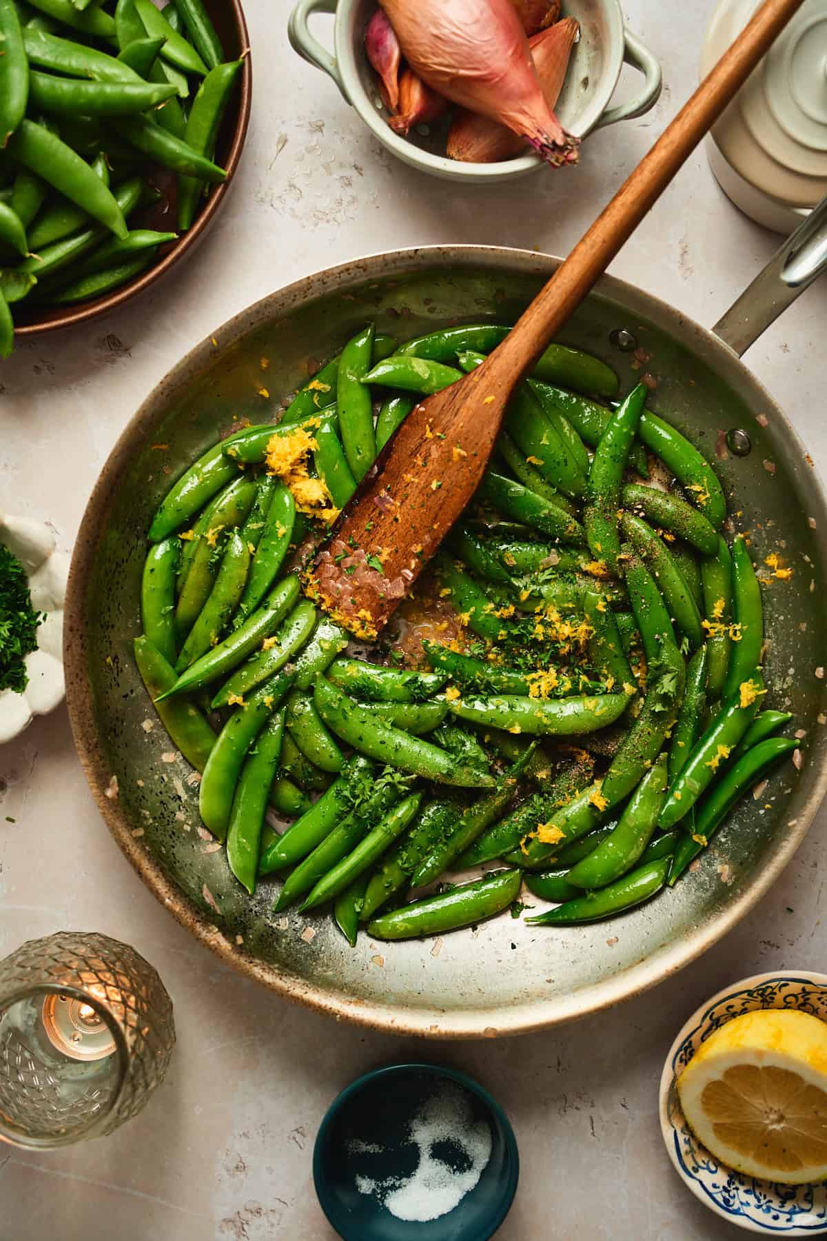 Lemon zest being stirred into snap peas in a skillet with a wooden spoon.