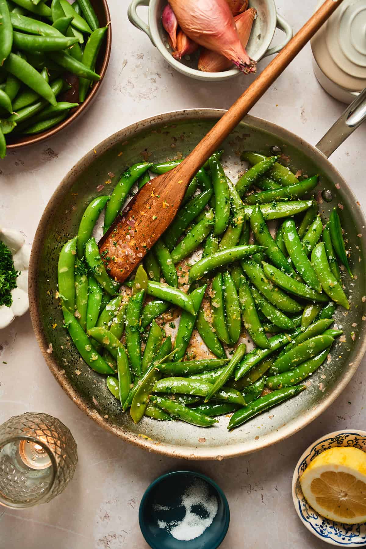 Snap peas with lemon zest and parsley tossed together in a skillet with a wooden spoon.