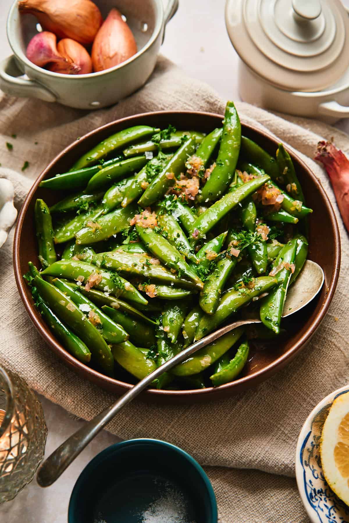 Overhead shot of buttery snap peas with parsley in a brown bowl, shallots and lemon in the background.