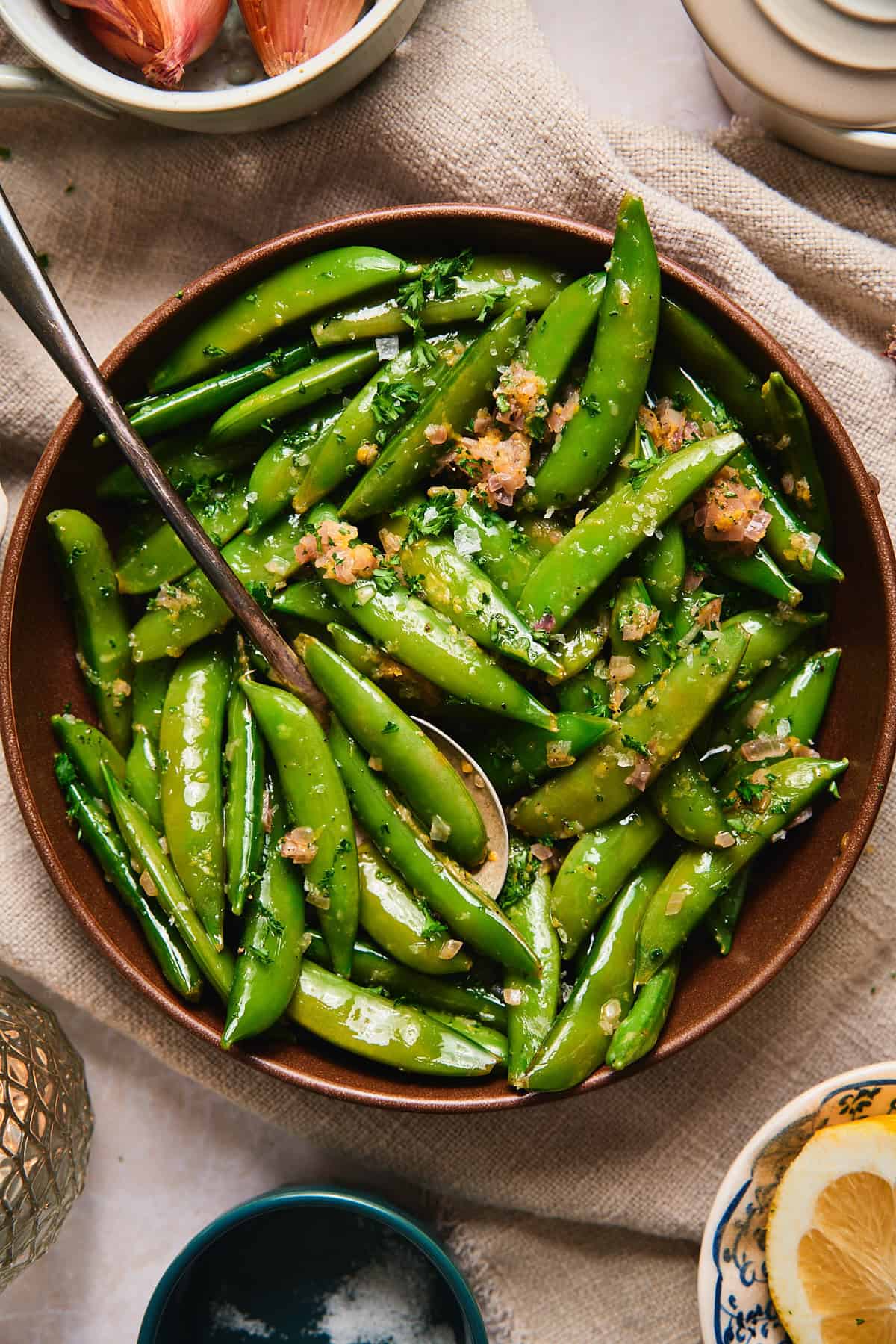 Flat lay of sautéed sugar snap peas in a brown bowl surrounded by shallots, lemon, salt, and a candle.