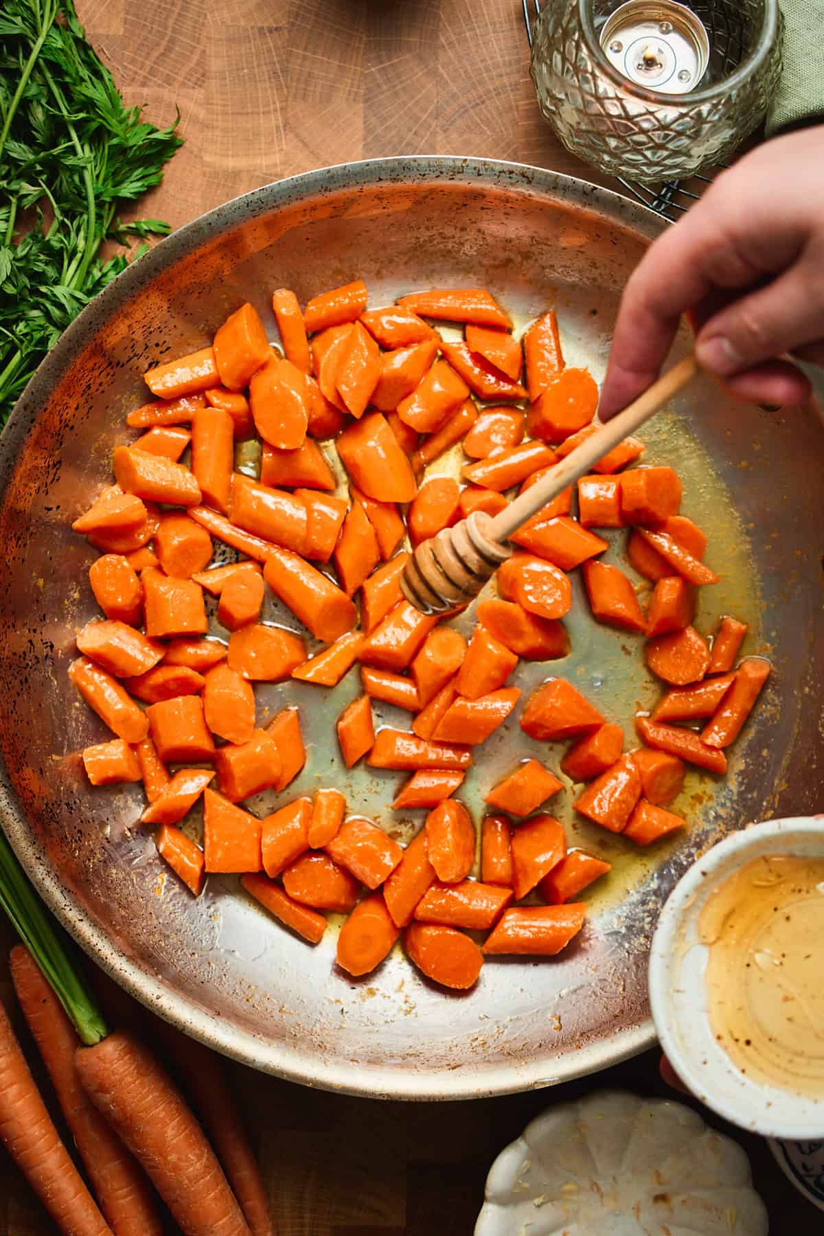 Hand drizzling honey over butter-coated carrots in a skillet with a honey dipper.